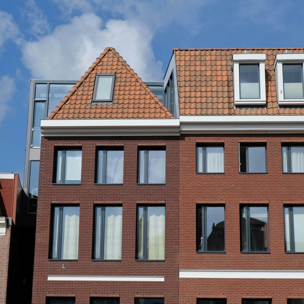 Red-brick residential building with large windows and terracotta-tiled pitched roofs under a blue sky.